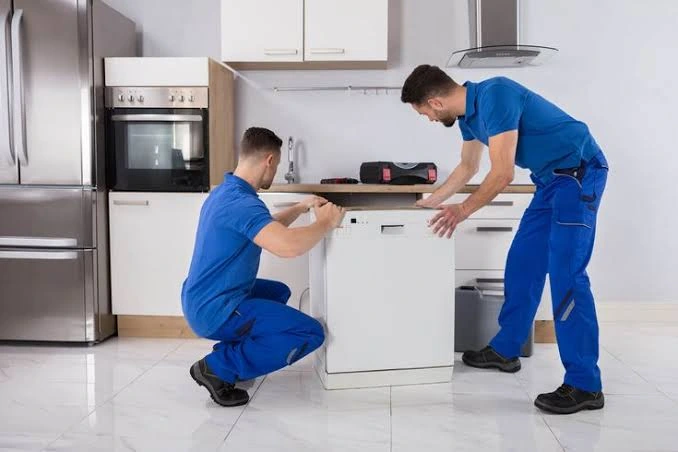 Two technicians positioning a dishwasher for installation in a kitchen